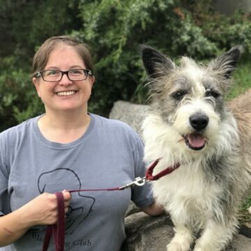 A person with short hair and glasses stands beside a short, long, wiry-coated dog.